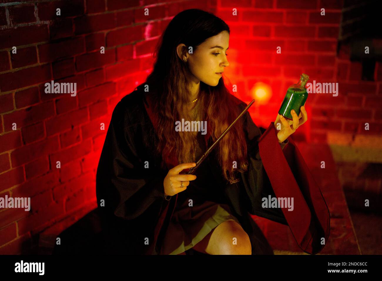 Junge Frau in Bademänteln sitzt mit Zauberstab und Zaubertrank Flasche Magisches Licht aus Zauberstab | Rot | Moody Stockfoto