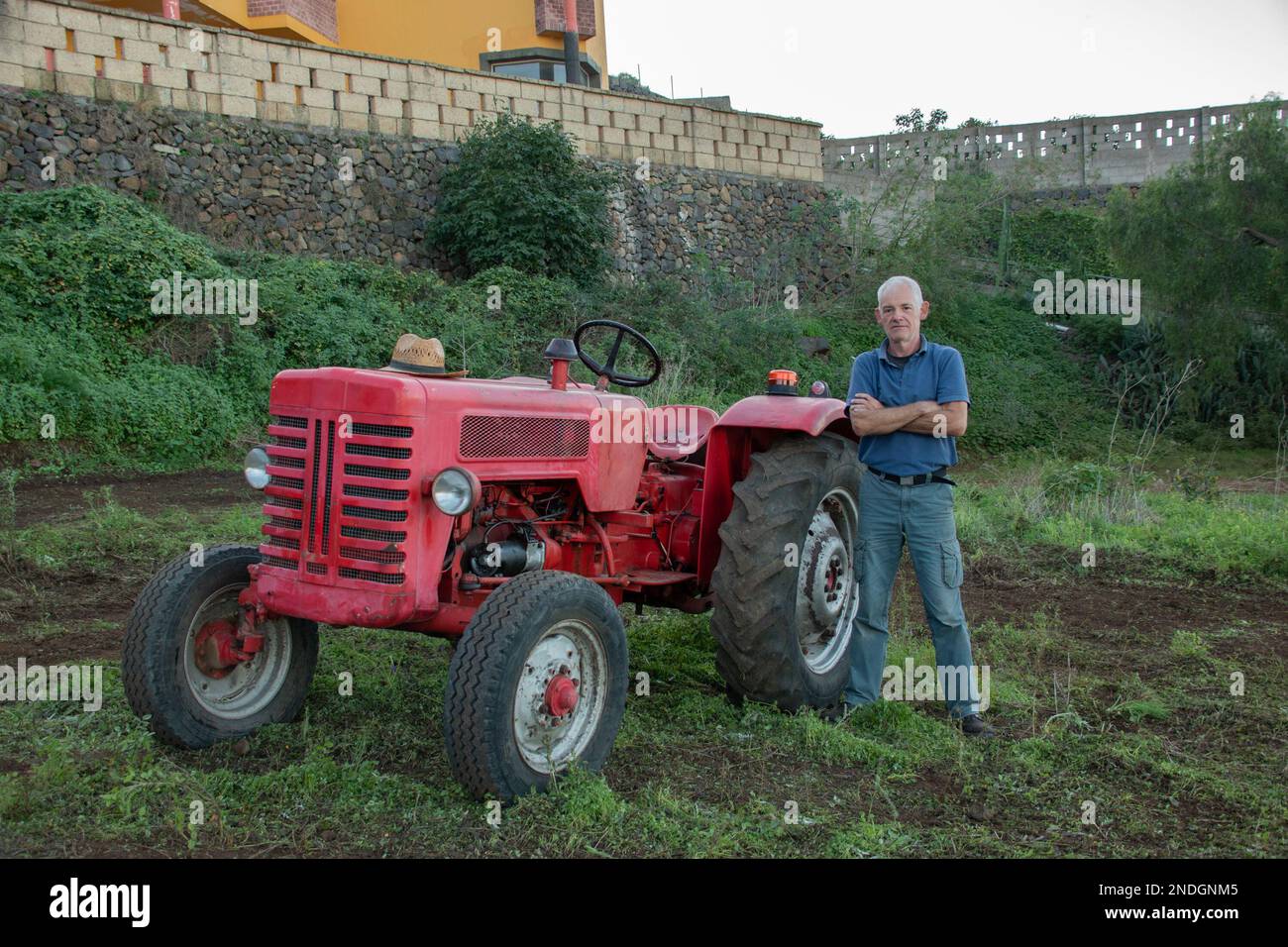 Die ganze Länge eines weißen Landwirts mit gekreuzten Armen und seinem Traktor auf seinem Ackerland Stockfoto
