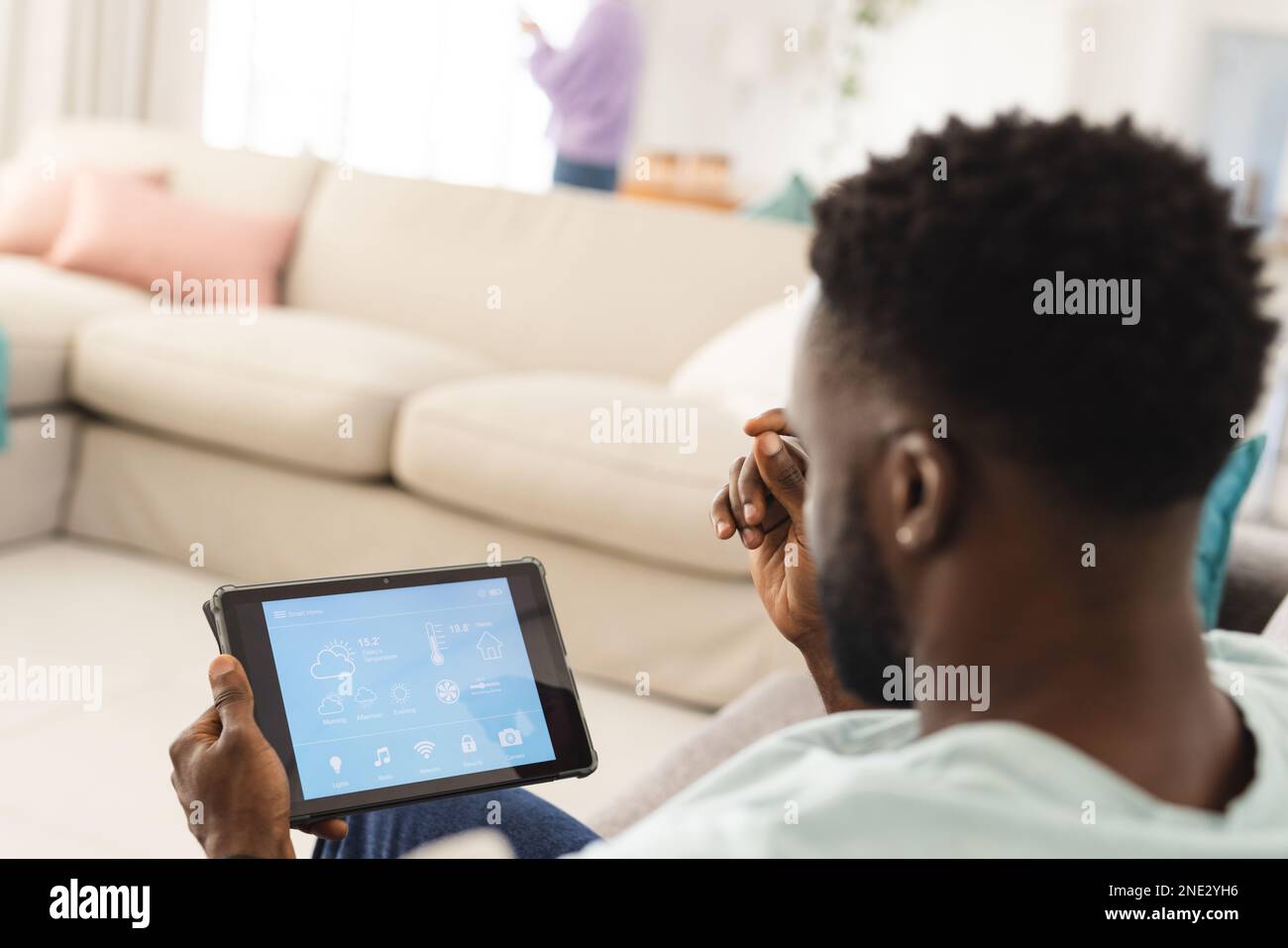 afroamerikanischer Mann, der auf dem Sofa im Wohnzimmer sitzt und ein Tablet mit Smart-Home-Schnittstelle verwendet. Verbringen Sie Zeit zu Hause, zu Hause und in Ihrem Leben Stockfoto