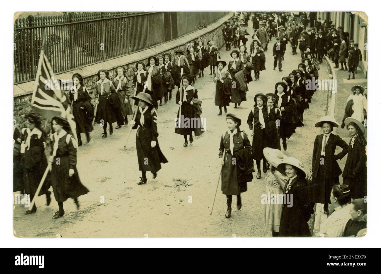 Originale patriotische Truppen aus Führerinnen und Pfadfinderinnen, die durch eine Stadt marschieren, mit einer Union-Jack-Flagge marschieren, eine öffentliche Kundschafter-Rallye, einige der Zuschauer tragen ein Reverskragen-Band, ca. 1912, Großbritannien Stockfoto