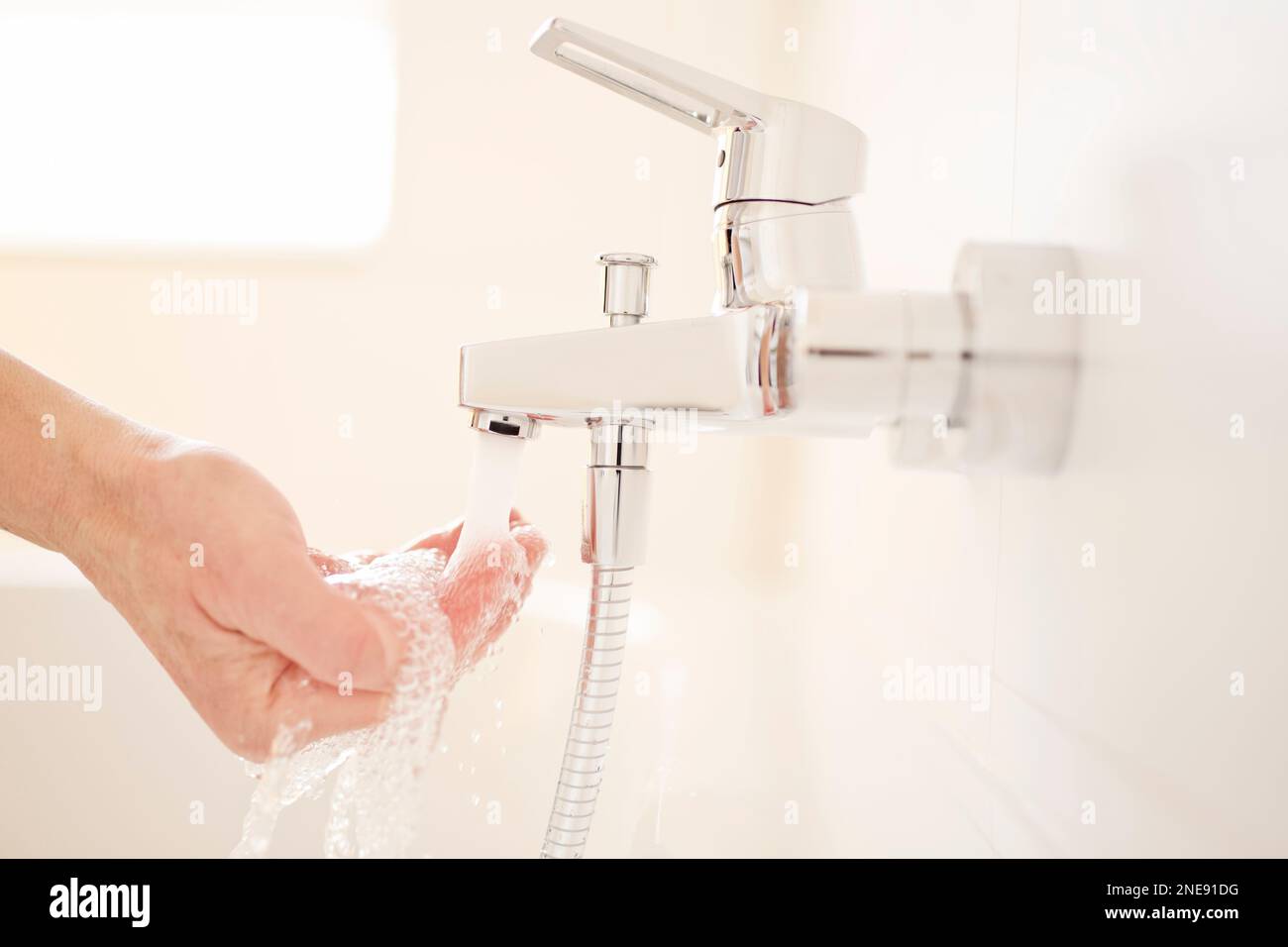 Männliche Hand prüft die Temperatur des fließenden Wassers aus einem Wasserhahn in einem hellen Badezimmer- selektiver Fokus Stockfoto