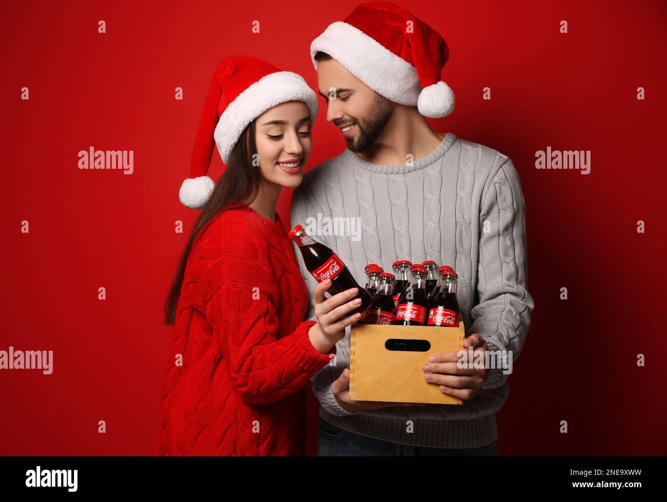 MYKOLAIV, UKRAINE - 27. JANUAR 2021: Junges Paar in Weihnachtshüten, das eine Kiste mit Flaschen Coca-Cola auf rotem Hintergrund hält Stockfoto