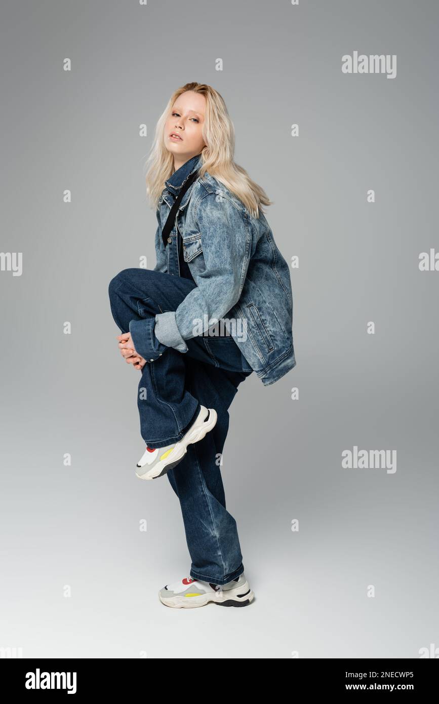 Eine lange, blonde Frau in stilvollem Denim-Outfit und Sneakers, die auf einem Bein auf einem grauen Stock-Bild posieren Stockfoto