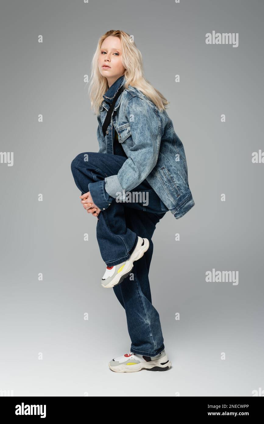 Eine blonde Frau in trendigen Jeans-Outfits und Sneakers, die auf einem Bein auf einem grauen Stock-Bild posieren Stockfoto