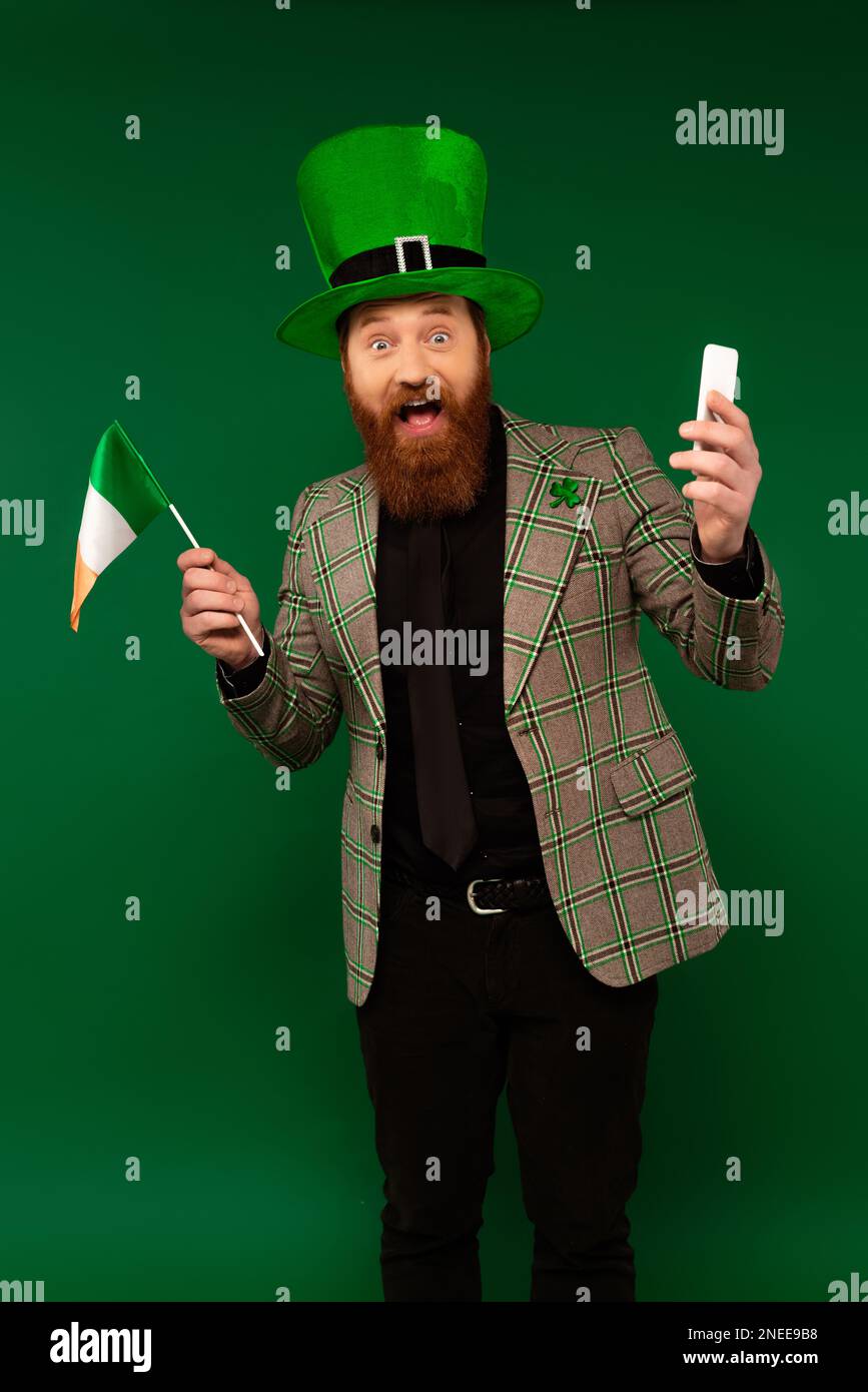 Aufgeregter Mann mit Hut und Klee mit Smartphone und irischer Flagge isoliert auf grünem Stockbild Stockfoto