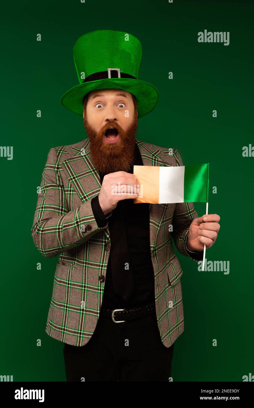 Ein schockierter bärtiger Mann mit Hut, der die irische Flagge hält, isoliert auf einem grünen Stock-Image Stockfoto