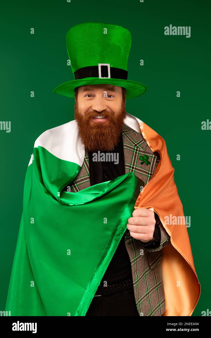 Lächelnder Mann mit Hut und Klee, eingewickelt in irischer Flagge, isoliert auf grünem Stockbild Stockfoto