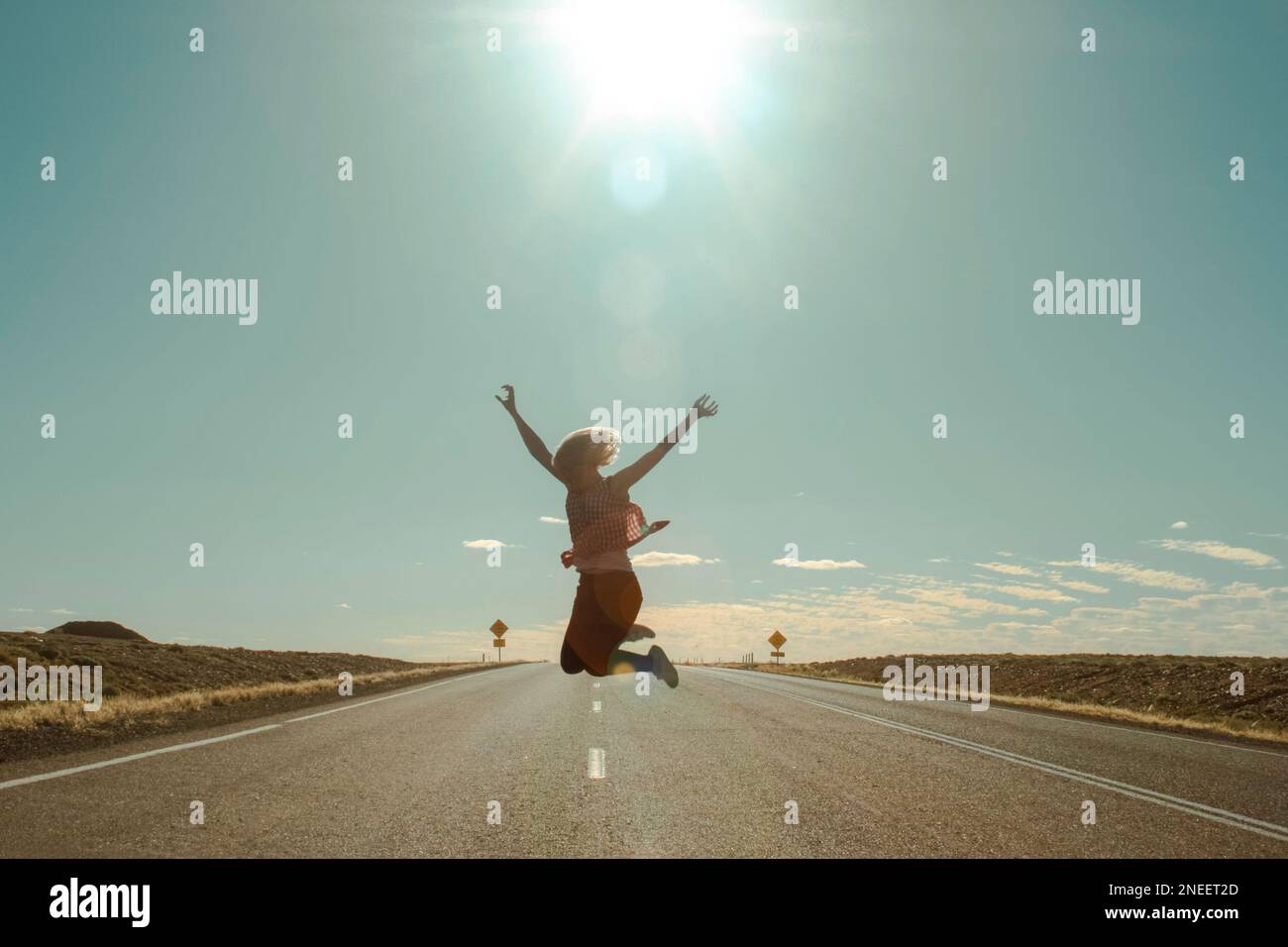 Silhouette einer glücklichen jungen erwachsenen Frau, die auf einer leeren Straße in die Luft springt, mit Sonne und Horizont im Hintergrund und Linsenreflexionen Stockfoto