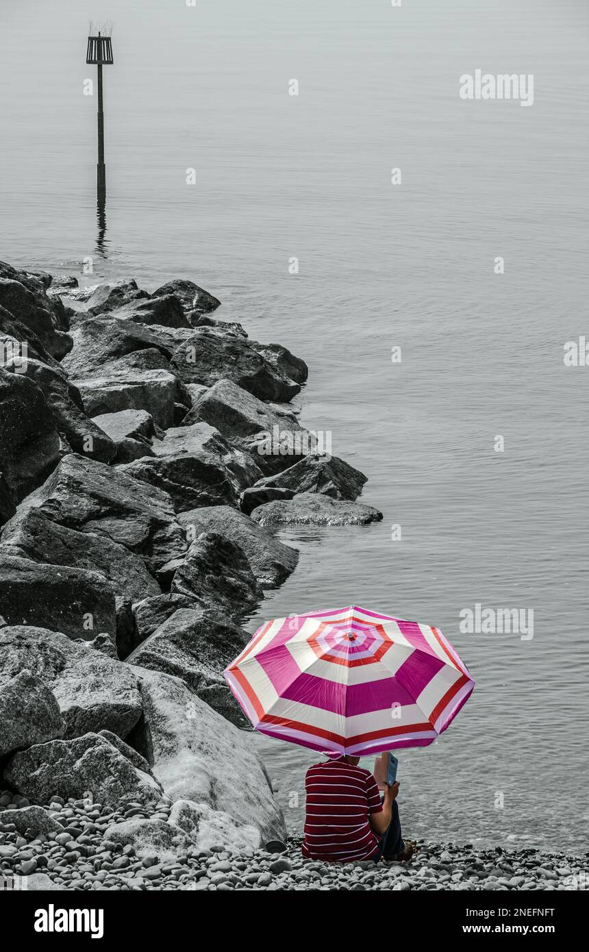 Eine Person, die ein Buch unter einem rosa Sonnenschirm in Sidmouth Beach, Devon, England liest Stockfoto