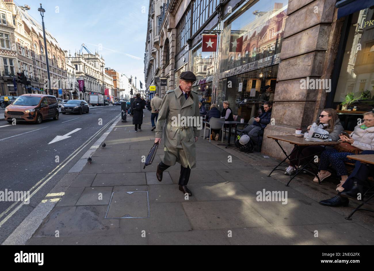 Ein Mann mit Regenmantel und flacher Mütze macht sich auf den Weg nach Piccadilly im Zentrum Londons, während die Leute draußen sitzen, England, Großbritannien Stockfoto