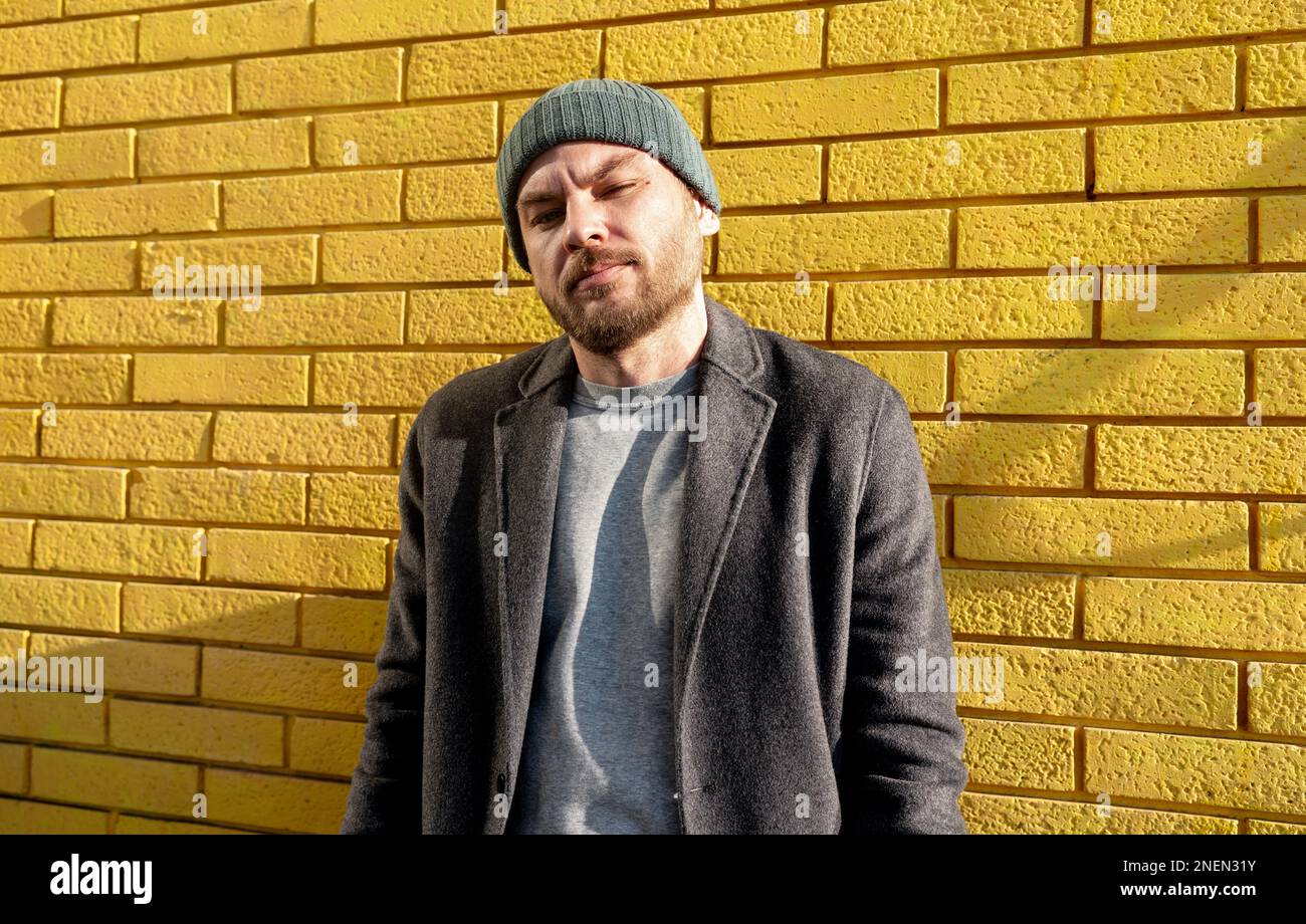 Stilvoller, erwachsener Mann mit Hut und Mantel, der an einer gelben Backsteinwand steht und in die Kamera schaut. Stockfoto