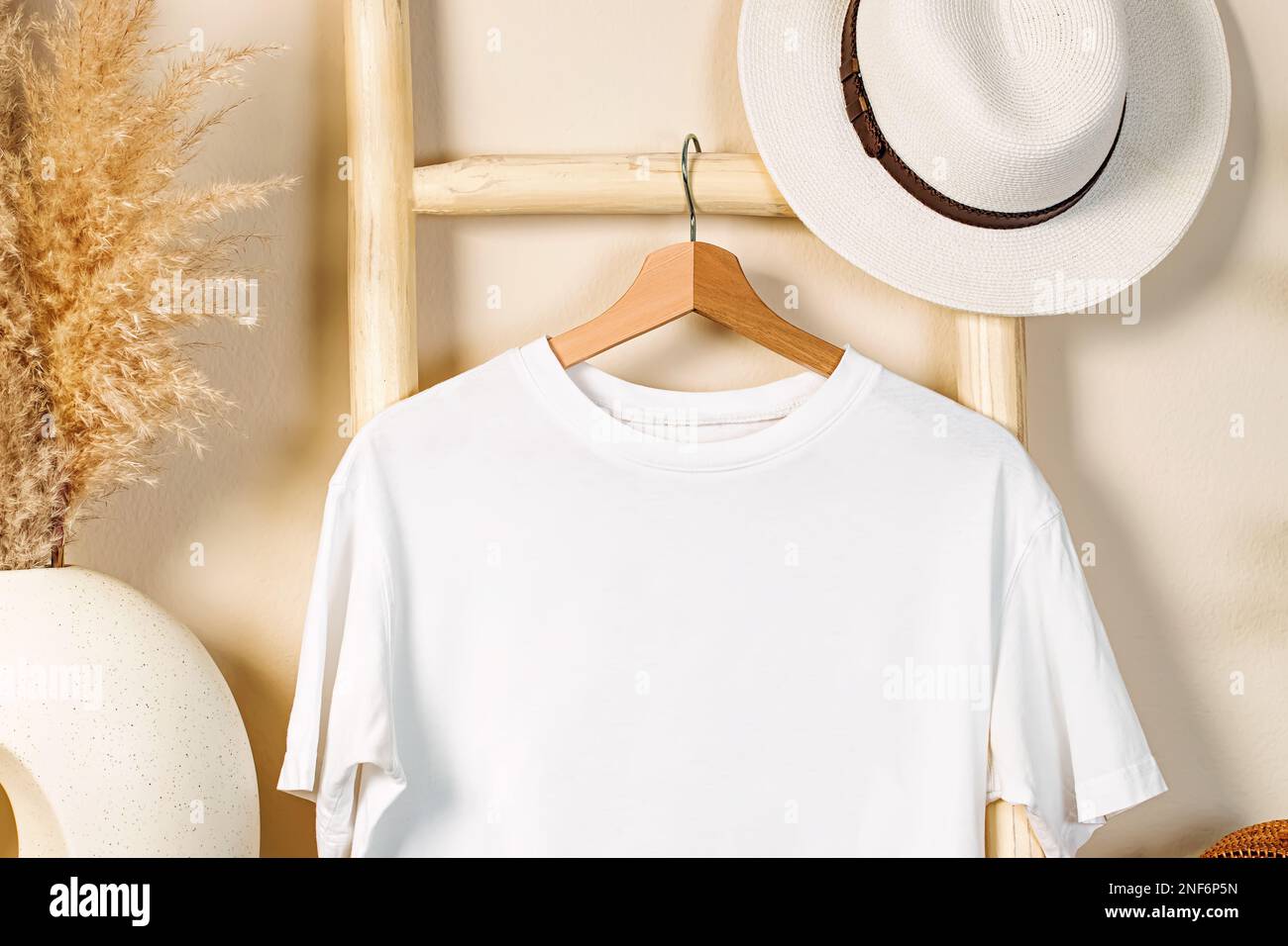 Weißes T-Shirt auf dem Holzbügel, Hut und trockenes Gras, T-Shirt-Mock im Bo-Interieur Stockfoto
