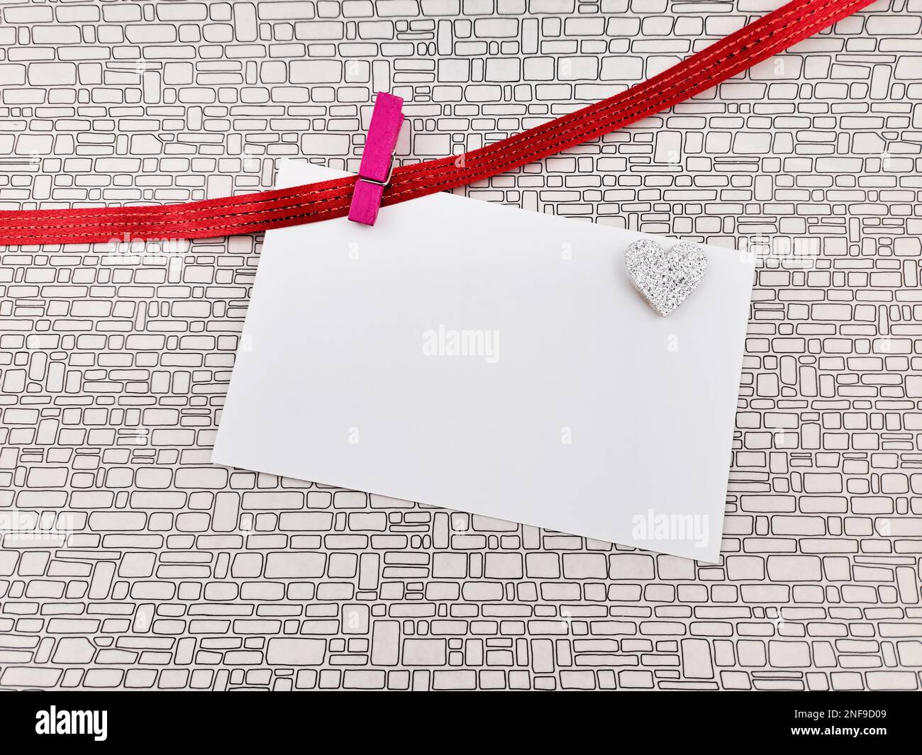 Weiße Valentinstagskarte, die an einer roten Schnur mit Wäscheklammer hängt, Hintergrund mit Mauermuster. Stockfoto Weiße Valentinstagskarte, die an einer roten Schnur mit Wäscheklammer hängt, Hintergrund mit Mauermuster. Stockfoto