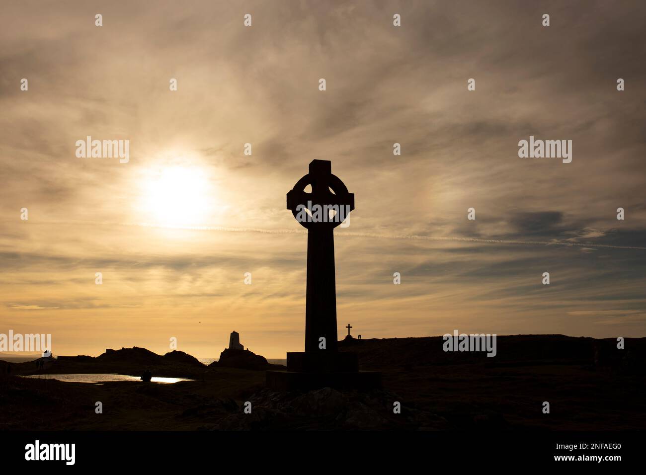 Silhouette eines keltischen Kreuzes mit der Abendsonne direkt dahinter. Mit einem Leuchtturm, Gebäuden und einem Kreuz im Hintergrund Stockfoto