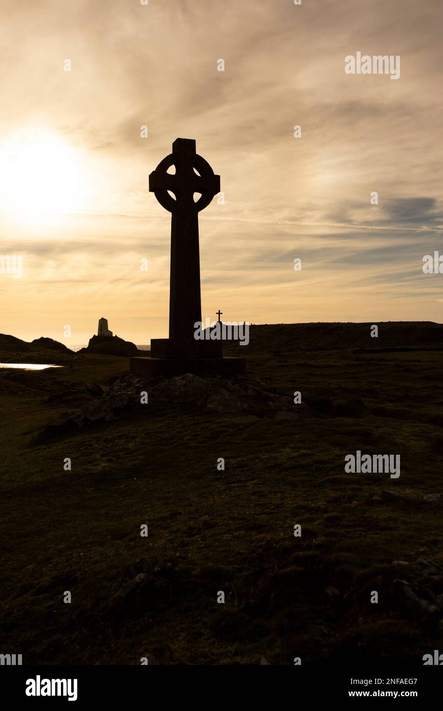 Silhouette eines keltischen Kreuzes mit der Abendsonne direkt dahinter. Mit einem Leuchtturm, Gebäuden und einem Kreuz im Hintergrund Stockfoto