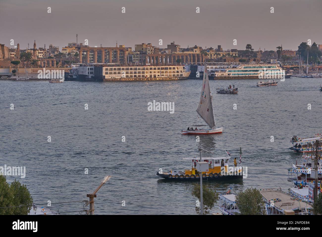 Luxor, ägyptischer Sonnenuntergang vom Westufer aus, der den Nil mit Felukken, Kreuzfahrtschiffen und dem Luxor Tempel am Ostufer zeigt Stockfoto