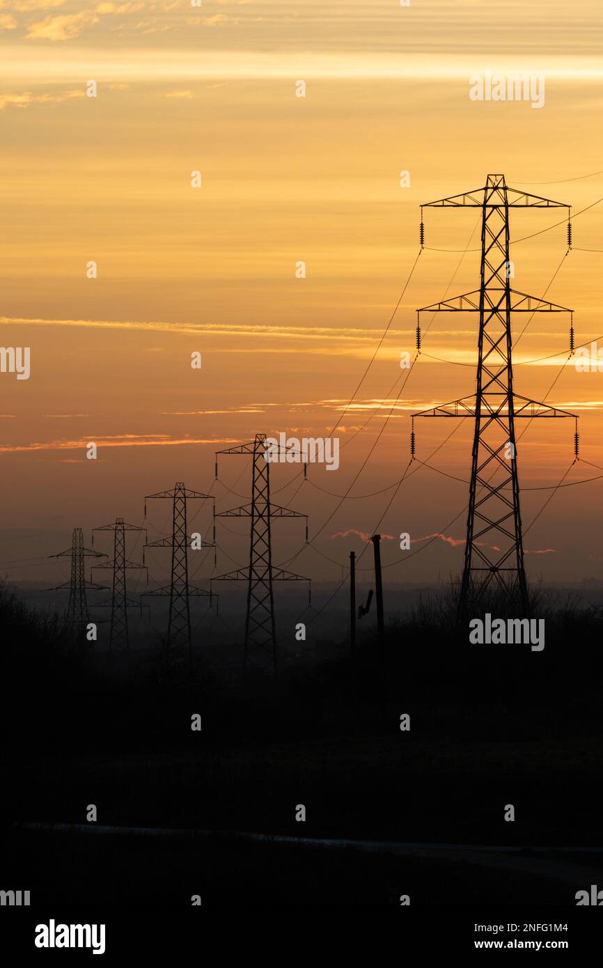 Silhouette von Strommasten vor dem orangefarbenen Himmel bei Sonnenuntergang, Großbritannien. Thema: Energieverbrauch, Strom, Rechnungen, steigende Kosten, Stromnetz Stockfoto