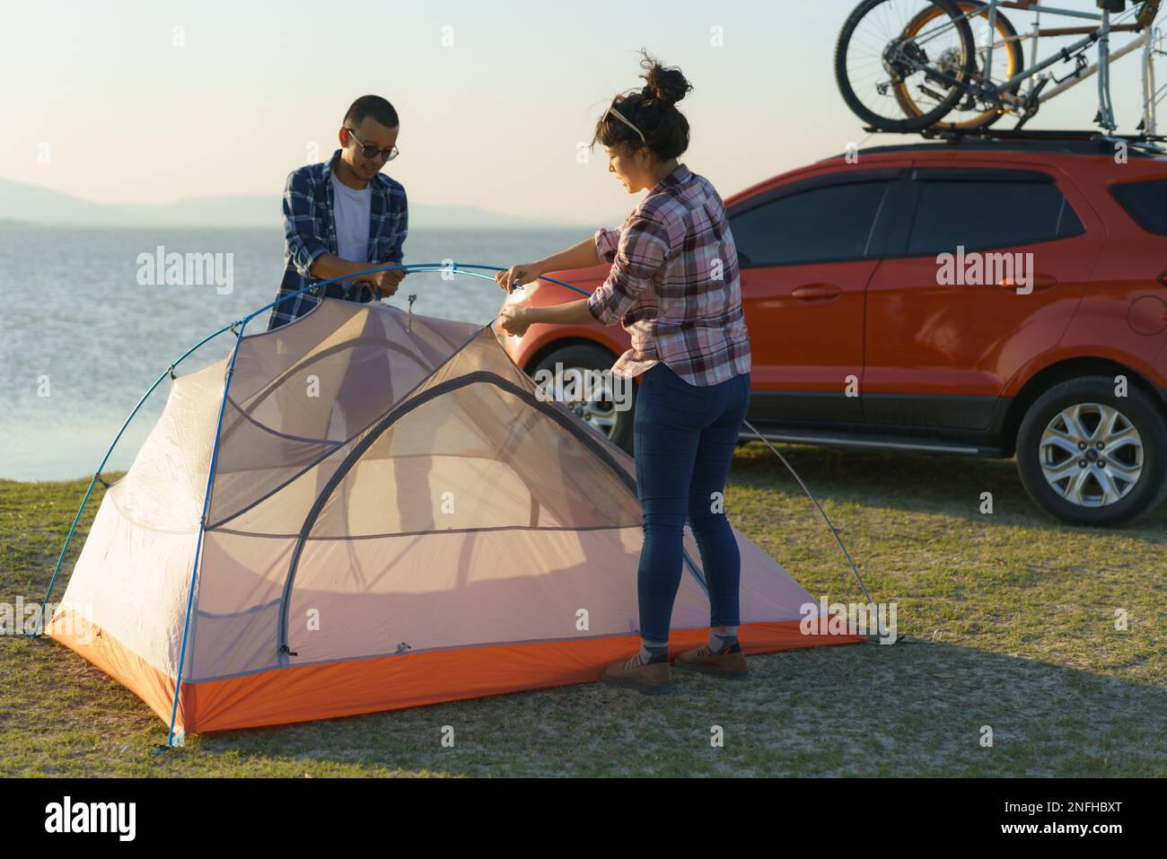 Ein asiatisches Paar bereitet ein Zelt vor, um auf dem Rasen zu campen, während der See im Hintergrund bei Sonnenuntergang liegt Stockfoto