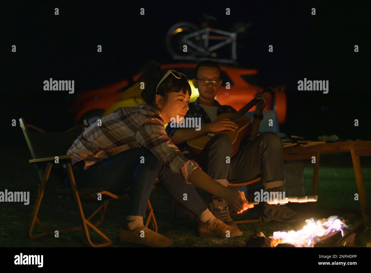 Asiatische Frau, die Marshmallows anstoßt, während ihr Freund am Lagerfeuer Gitarre spielt, wo sie ihre Zelte für das Camping am See nachts aufstellen. Stockfoto