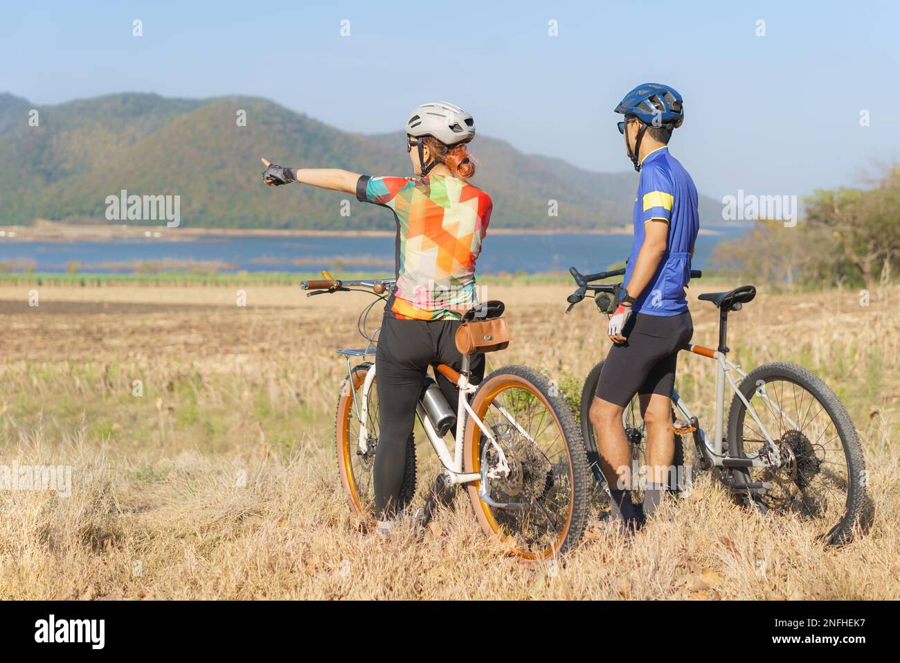 Asiatisches Fahrradpaar, das den Blick auf den See und die Berge genießt und sich unterhält, während es eine Pause von der morgendlichen Radtour macht Stockfoto