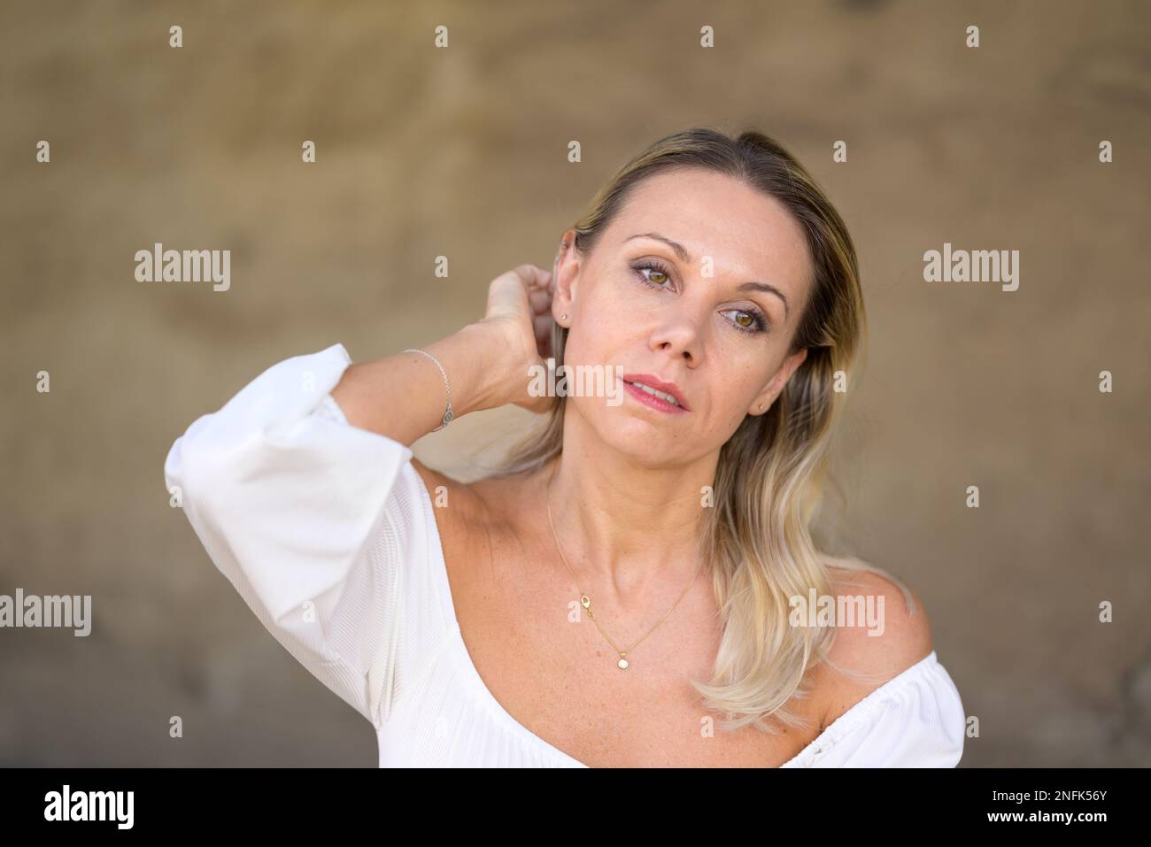 Das Porträt einer blonden Frau mittleren Alters mit einer weißen Bluse führt eine Hand durch ihr Haar mit einem durchdachten Gesichtsausdruck vor einer braunen Wand Stockfoto