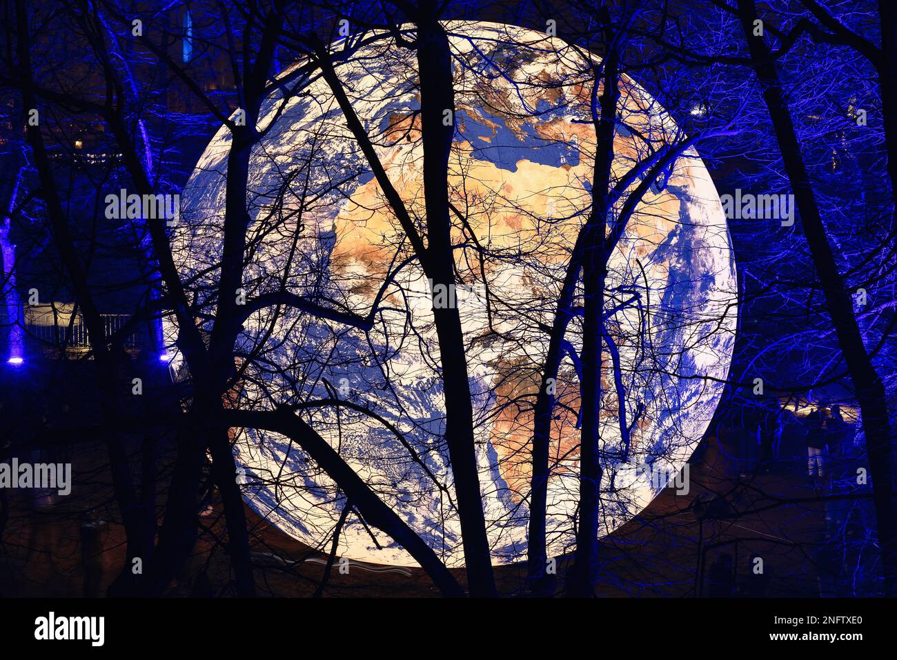 Brescia - Italia. 15. Februar 2023: Light is Life (Festa delle Luci A2A), „Floating Earth“ von Luke Jerram inmitten von blau beleuchteten, blattlosen Ästen von Bäumen Stockfoto