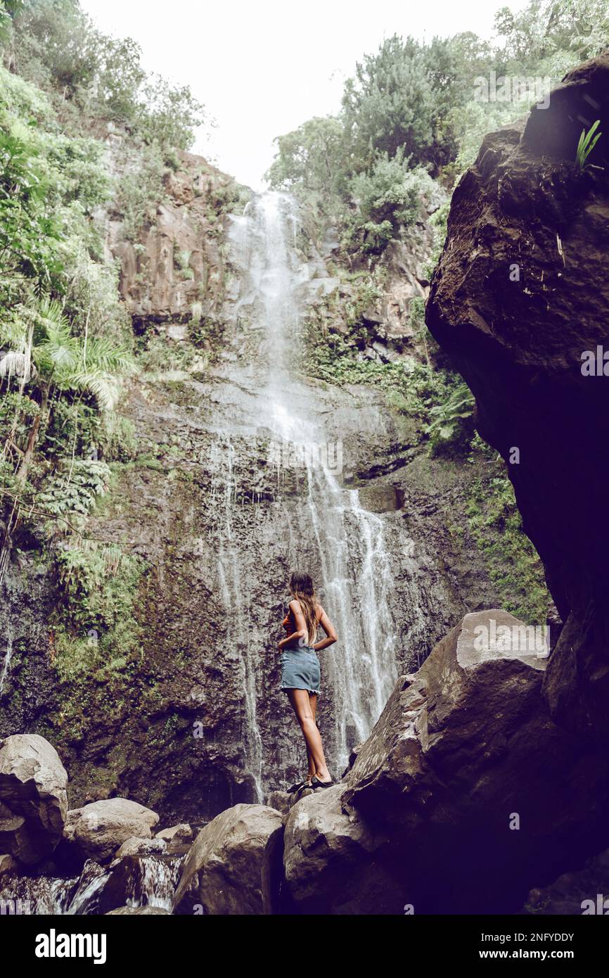Junge Frau, die auf einem Felsen steht und den Wasserfall beobachtet. Reisen und Abenteuer. Die Natur. Maui. Weg nach Hana Stockfoto