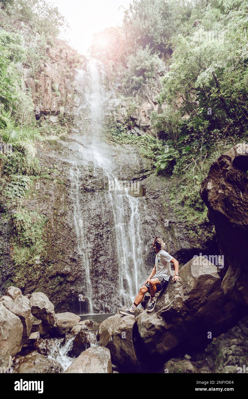 Junger Mann, der auf einem Felsen sitzt und den Wasserfall beobachtet. Reisen und Abenteuer. Die Natur. Maui. Weg nach Hana Stockfoto