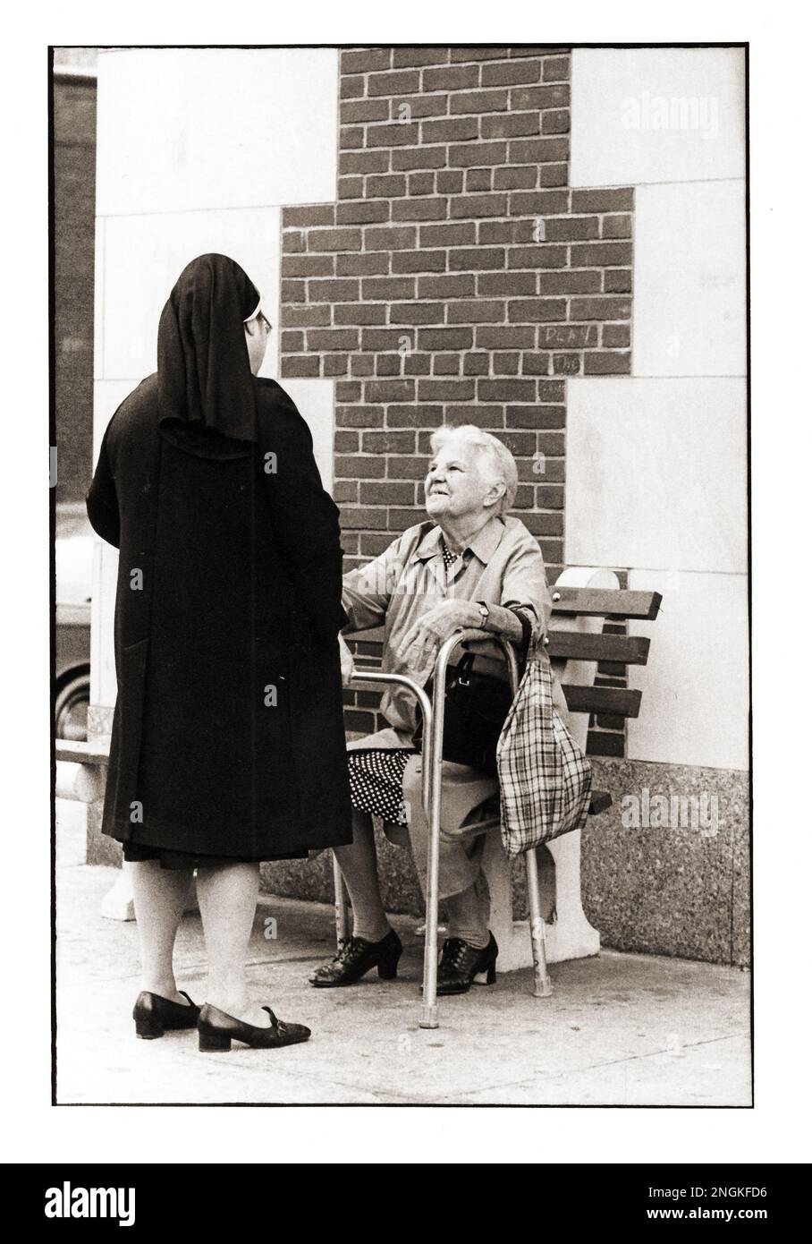Eine Nonne hält an, um sich mit einer älteren Frau vor einem Gebäude zu unterhalten, dessen Ziegel die Form eines Kruzifix bilden. Auf der Seventh Avenue in Park Slope, Brooklyn, New York. Etwa Ende 190s. Stockfoto