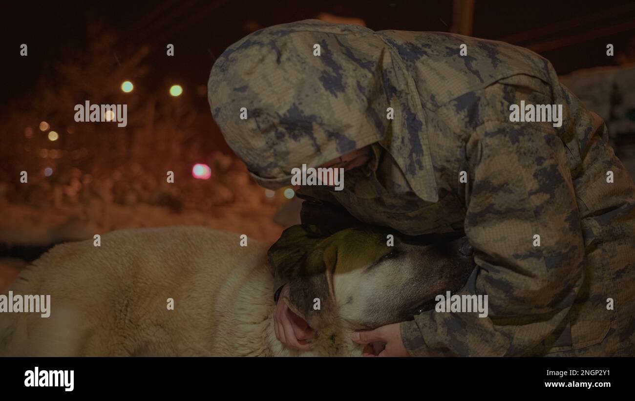 Militärisch gekleideter, bespitzelter junger Mann, der bei Schneewetter mit einem Sivas Kangal-Hund spielt Stockfoto