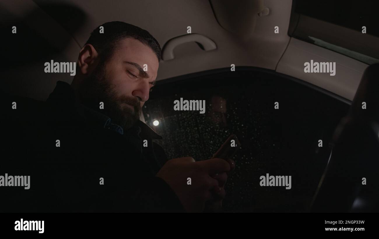 Der attraktive junge Mann vergisst den geschäftigen Tag, während er auf dem Beifahrersitz des Autos SMS schreibt Stockfoto