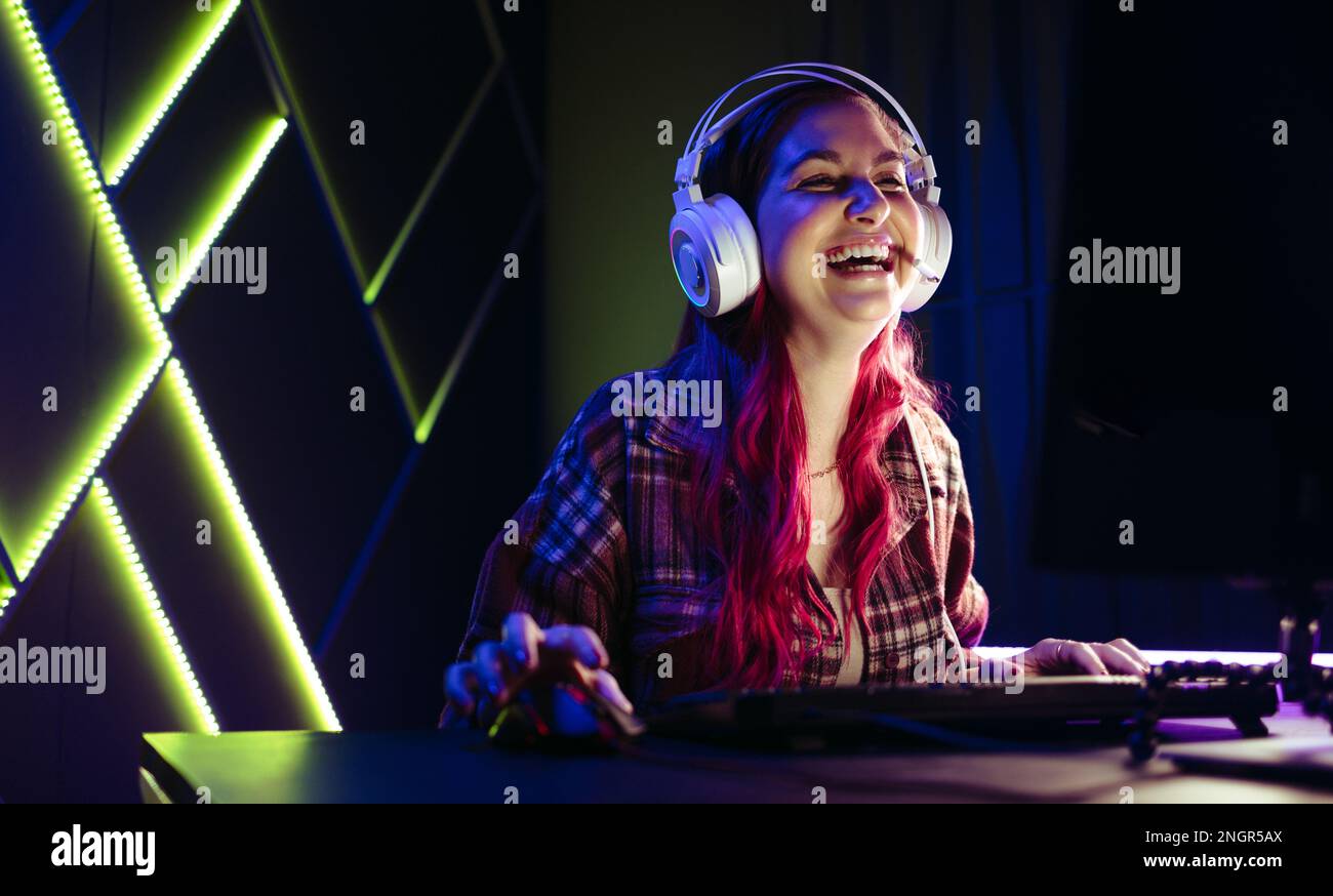 Eine Frau, die mit einem Headset an ihrer Gaming-Station sitzt und über ihren letzten Sieg in einem Online-Spiel lacht. Weibliche Gamer amüsieren sich als Stockfoto