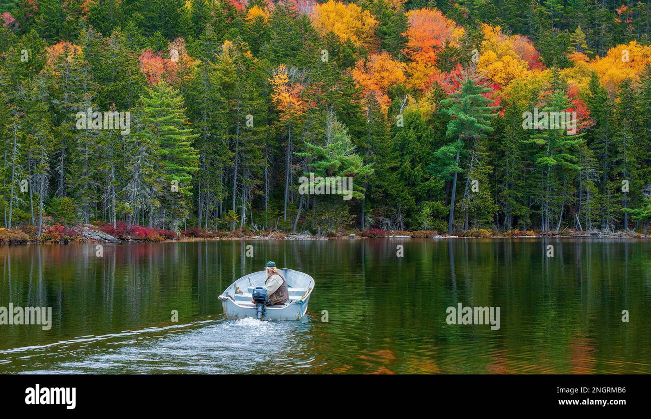 Ein Mann in seinen 60ern, der in einem Motorboot auf einem Bergsee unterwegs ist. Mischwald in Herbstfarben, die sich auf gerillten Gewässern widerspiegeln. Borestone Mountain, Maine. Stockfoto