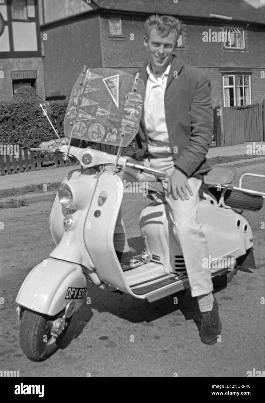 Ein modisch gekleideter junger Mann auf einem Lambretta Roller in Großbritannien Mitte der 1960er Jahre. Er trägt eine lässige Jacke, eine cremefarbene Hose mit Turnups und Wildlederschuhe. Orte, die der Roller besucht hat, werden als Wimpel auf der Windschutzscheibe angezeigt – darunter Paris und Oxford. Dieses klassische Foto aus der Zeit, als Roller zu einer „Mod“-Transportform mit Großbritanniens Jugendkultur wurden. Dieses Bild ist von einem alten Amateur-35mm-Negativ – ein klassisches 1960er-Foto. Stockfoto