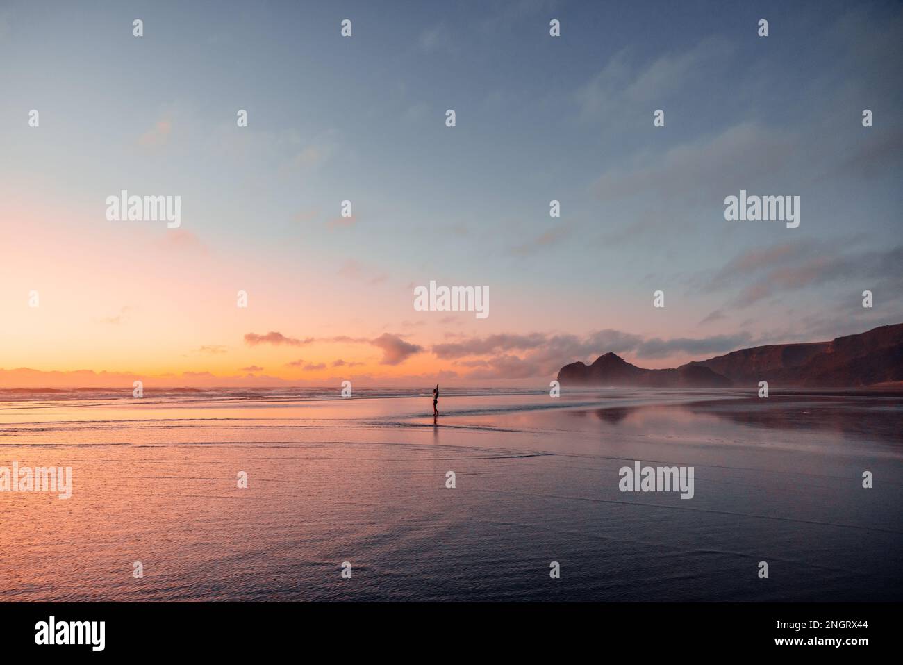 Eine junge Frau tanzt am Bethells Beach bei Sonnenuntergang, umgeben von leuchtenden Pink-, Violett- und Orangentönen. Stockfoto