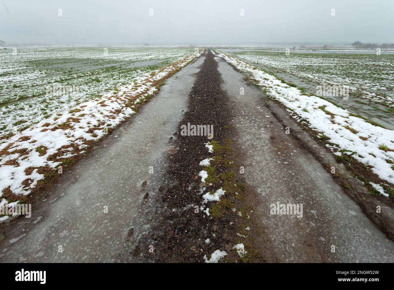 Eisige Schotterstraße durch Felder mit Schnee und Wasser, Blick an einem nebligen Tag, Czulczyce, Polen Stockfoto