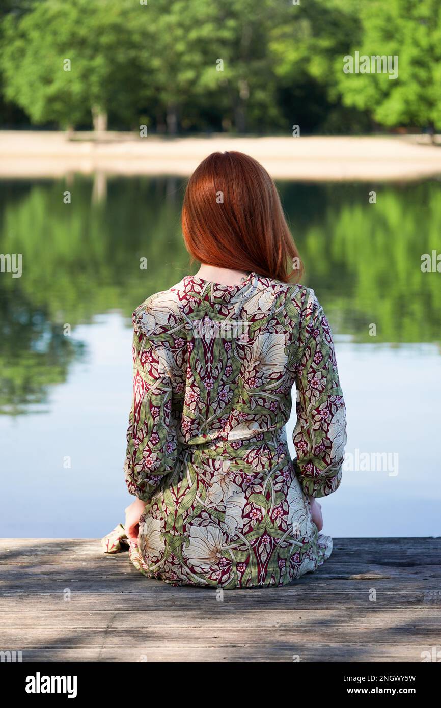 Rückansicht der jungen Frau Sommer Kleid tragen und sitzen auf hölzernen Pier auf idyllischen See Stockfoto