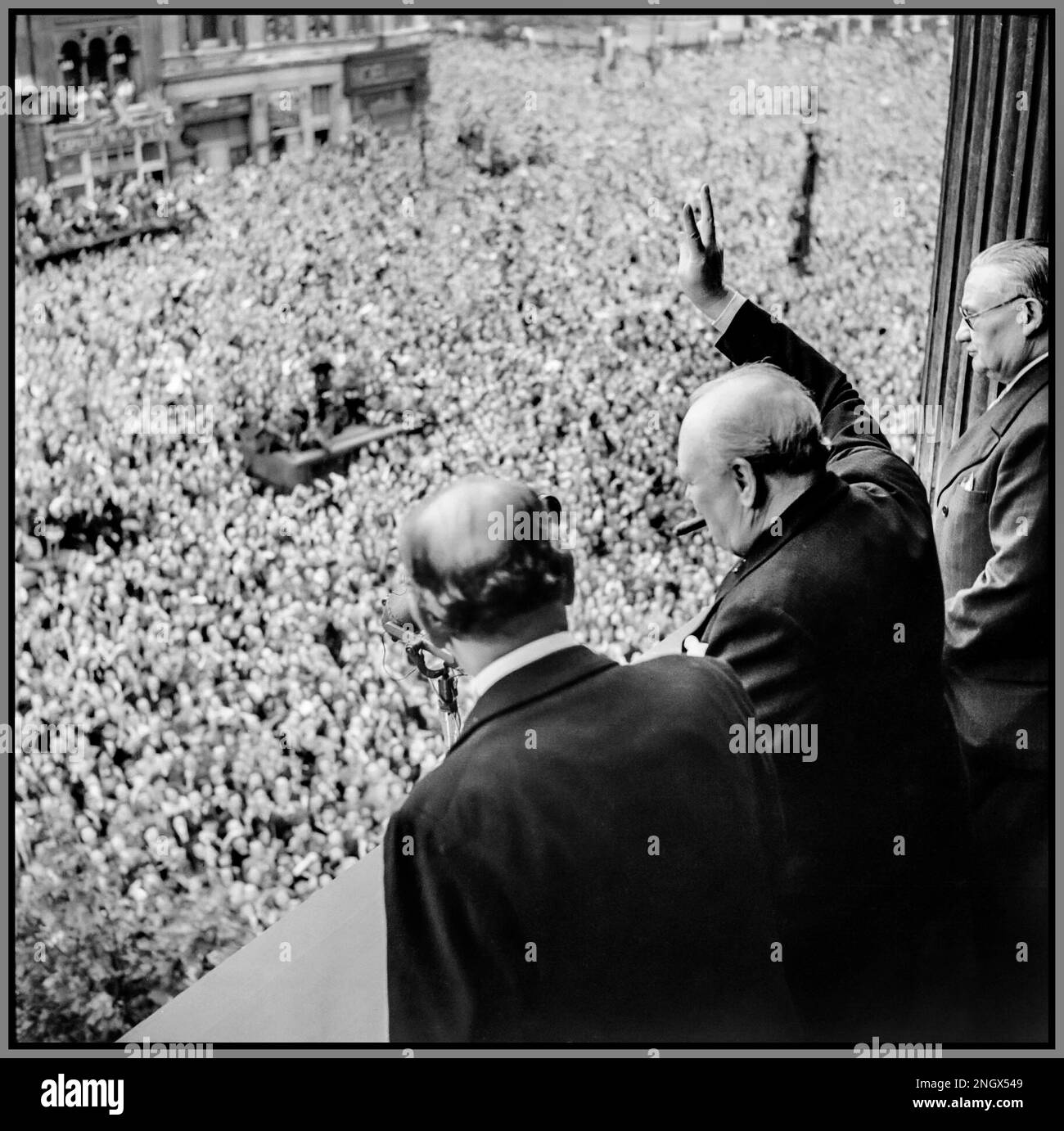 VE DAY WW2 (Sir) Winston Churchill winkt zu den ekstatischen Massen in Whitehall London, während sie VE Day, 8. Mai 1945, feiern. "Sieg in Europa" vom Londoner Balkon des Gesundheitsministeriums gibt Premierminister Winston Churchill mit seiner Markenzeichen Zigarre sein berühmtes "V for Victory" -Zeichen an massenhafte Menschenmassen in Whitehall am selben Tag, an dem er der britischen Nation übertrug, dass der Krieg mit Nazi-Deutschland gewonnen wurde, 8. Mai 1945 (VE Day). Zu Churchills Linken ist Sir John Anderson, der Schatzkanzler. Zu Churchills Rechten gehört Ernest Bevin, der Arbeitsminister. 8. Mai 1945 Stockfoto
