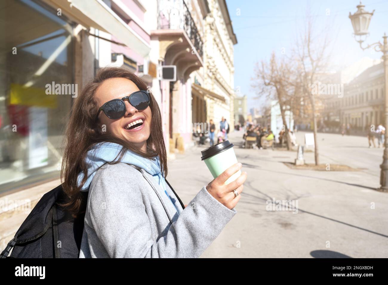 Zufriedene junge Frau mit Rucksack, die eine Kaffeetasse in der Hand hielt, während sie durch die Stadt spazierte. Stockfoto