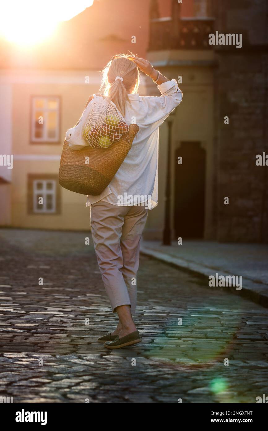 Frau mit wiederverwendbarer Tasche und Netztasche, die während des Sonnenuntergangs auf der Straße unterwegs ist. Nachhaltiger Lebensstil Stockfoto