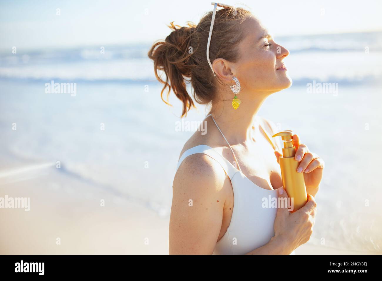 Glückliche, stilvolle Frau in weißer Strandbekleidung mit Sonnencreme am Strand. Stockfoto