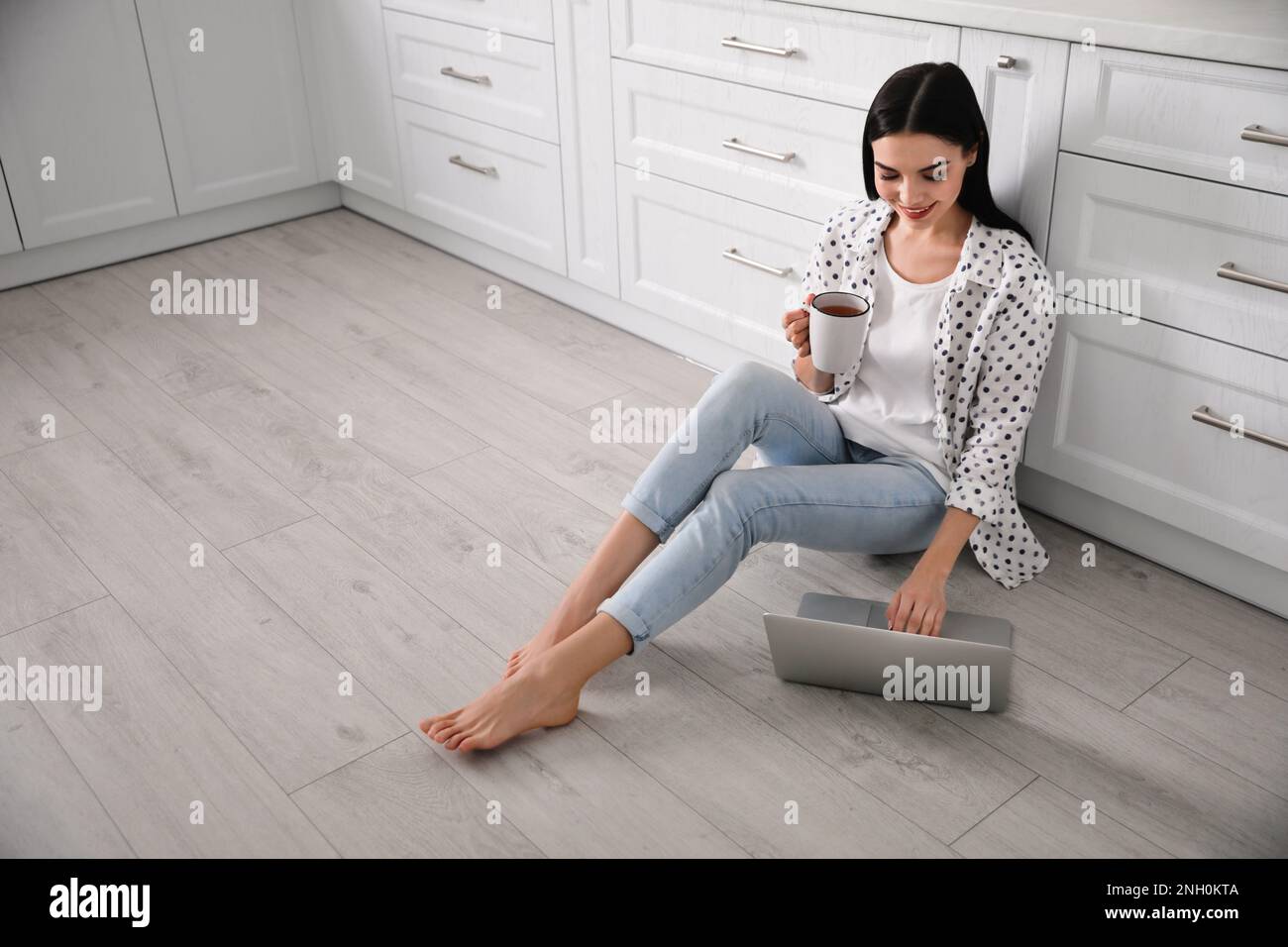 Glückliche Frau mit Laptop und einer Tasse Getränk, die auf dem warmen Boden in der Küche sitzt, Platz für Text. Heizung Stockfoto