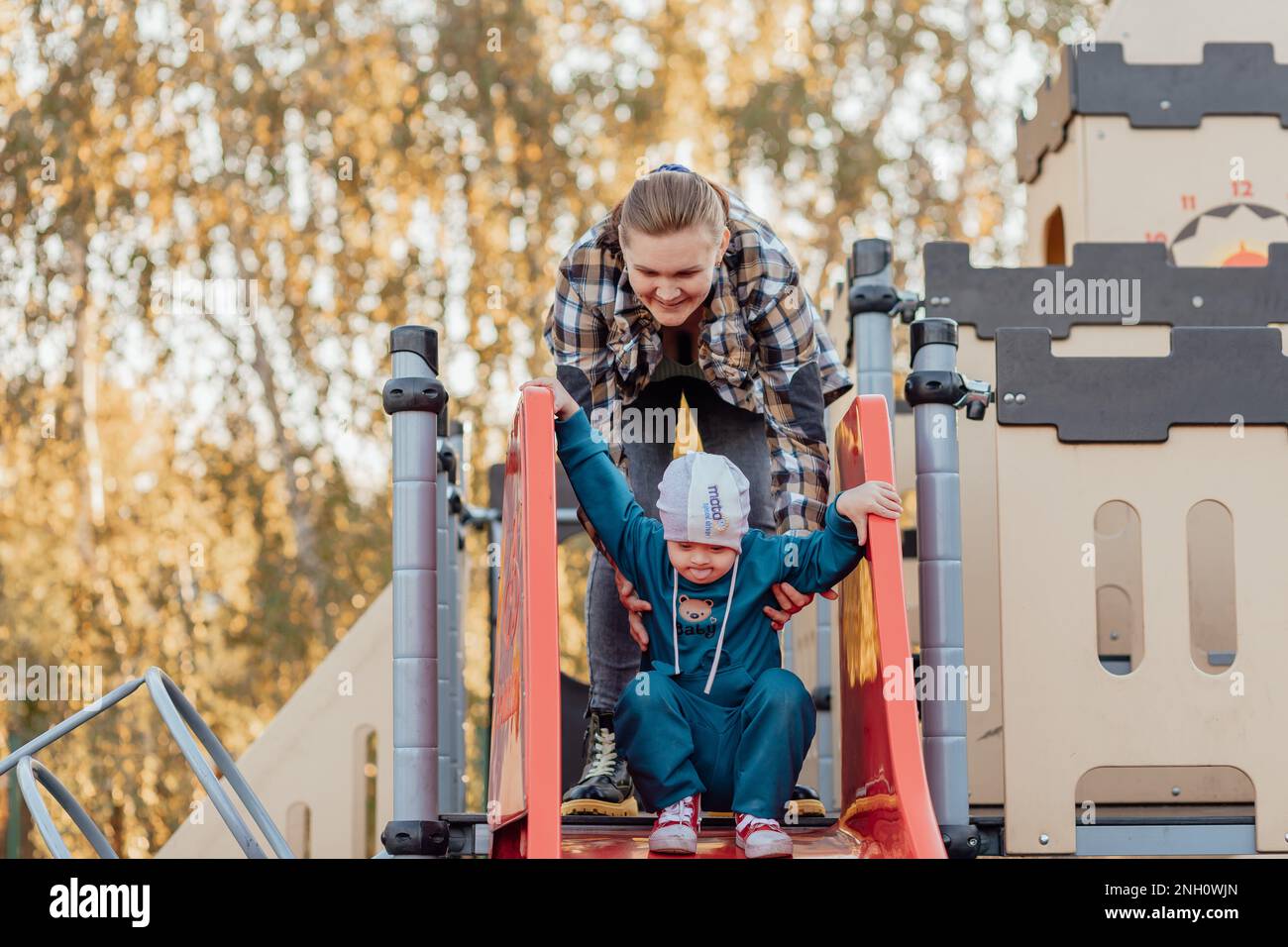 Ein Junge, eine Person mit Down-Syndrom, geht mit seiner Mutter durch den Park und geht die Kinderrutsche hinunter Stockfoto