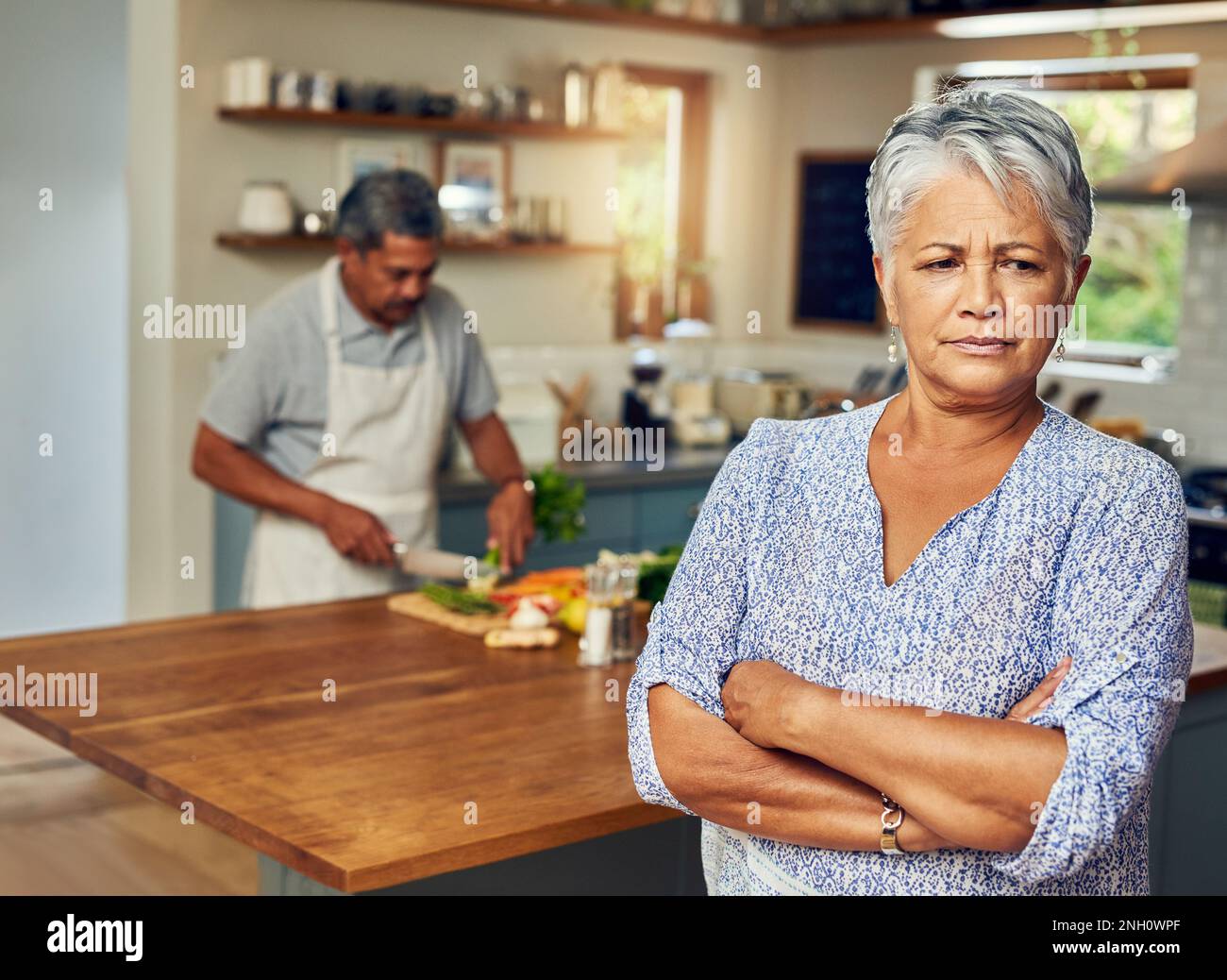 Er denkt, er sei ein besserer Koch als ich. Porträt einer erwachsenen Frau, die nach einem Streit mit ihrem Mann, der im Hintergrund kocht, aufgebracht wirkt. Stockfoto