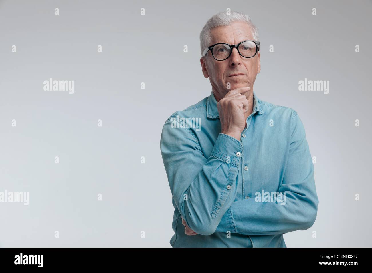 Porträt eines gutaussehenden Mannes in seinen 60s, der aufblickt und denkt, während er seine Arme vor grauem Hintergrund faltet Stockfoto