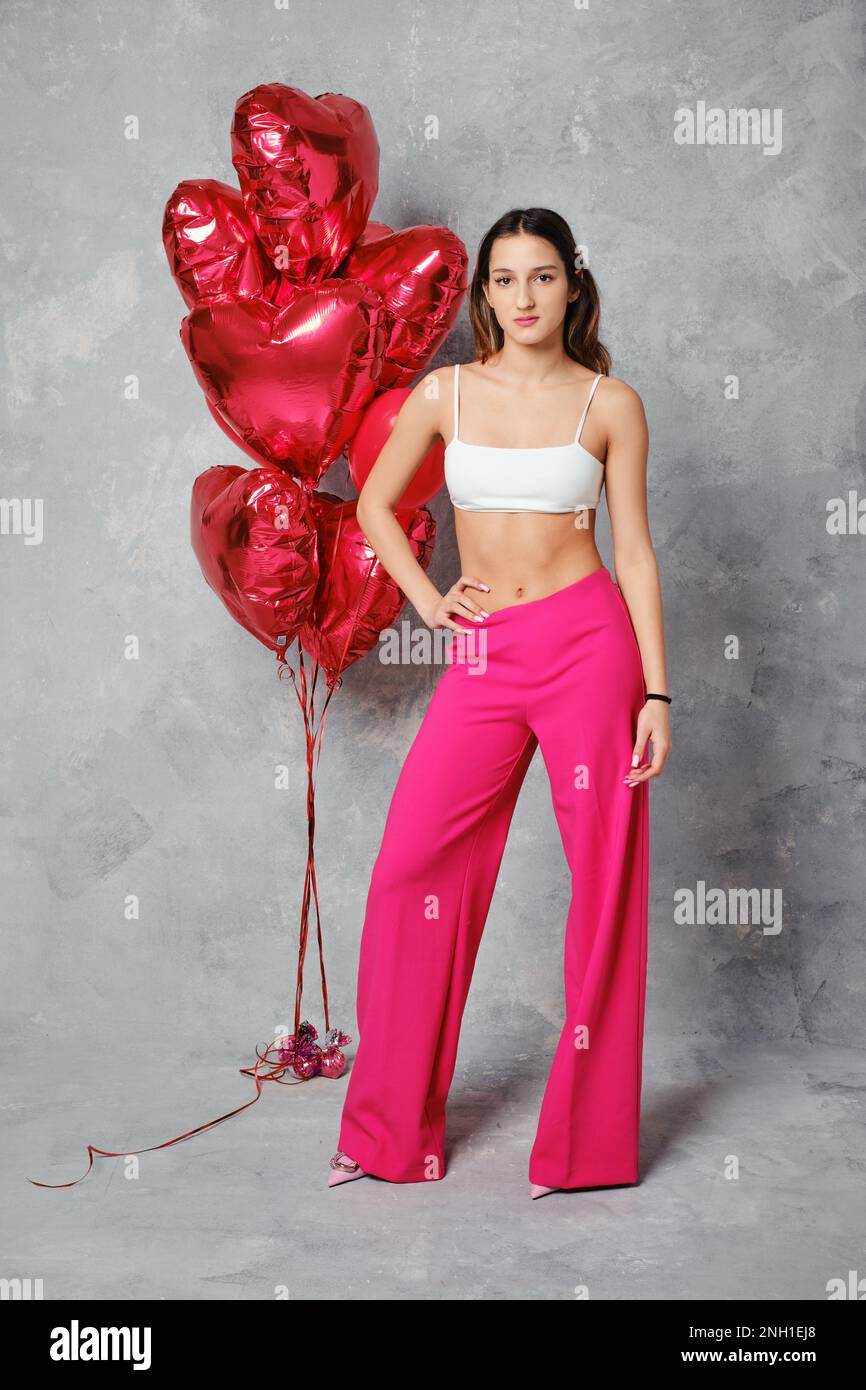 Fröhliche junge Frau in einer breiten, leuchtend pinkfarbenen Hose und weißem Schlauchoberteil, die neben Ballons im Studio posiert Stockfoto