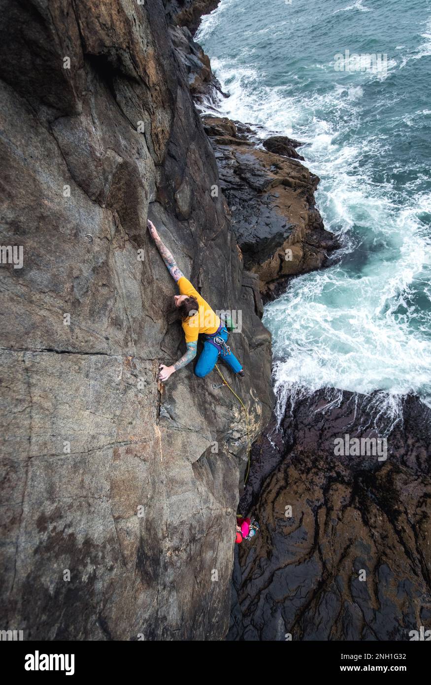 Ein Mann klettert auf eine Klippe am Meer und die Wellen schlagen darunter Stockfoto