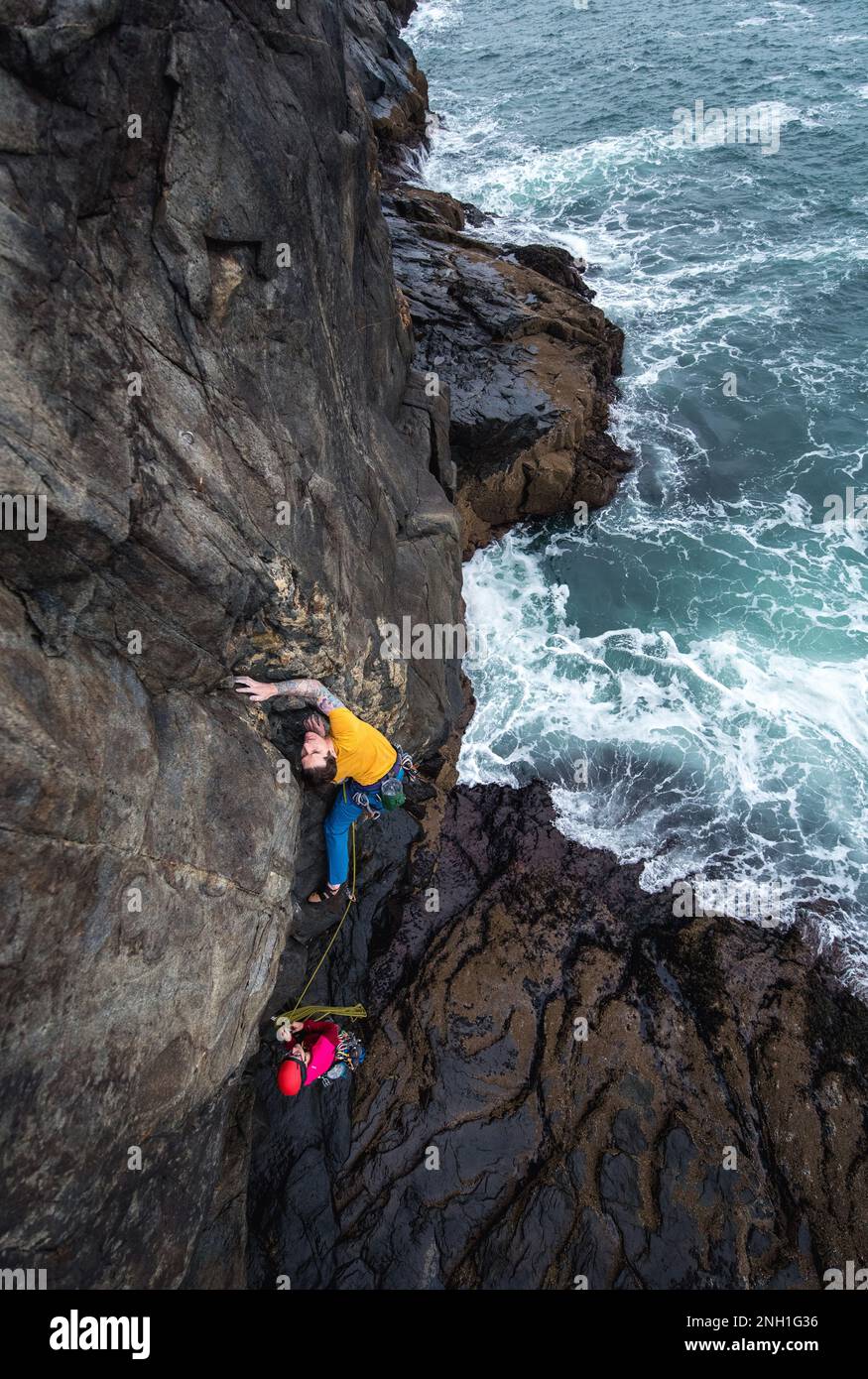 Ein Mann klettert auf eine Klippe am Meer und die Wellen schlagen darunter Stockfoto