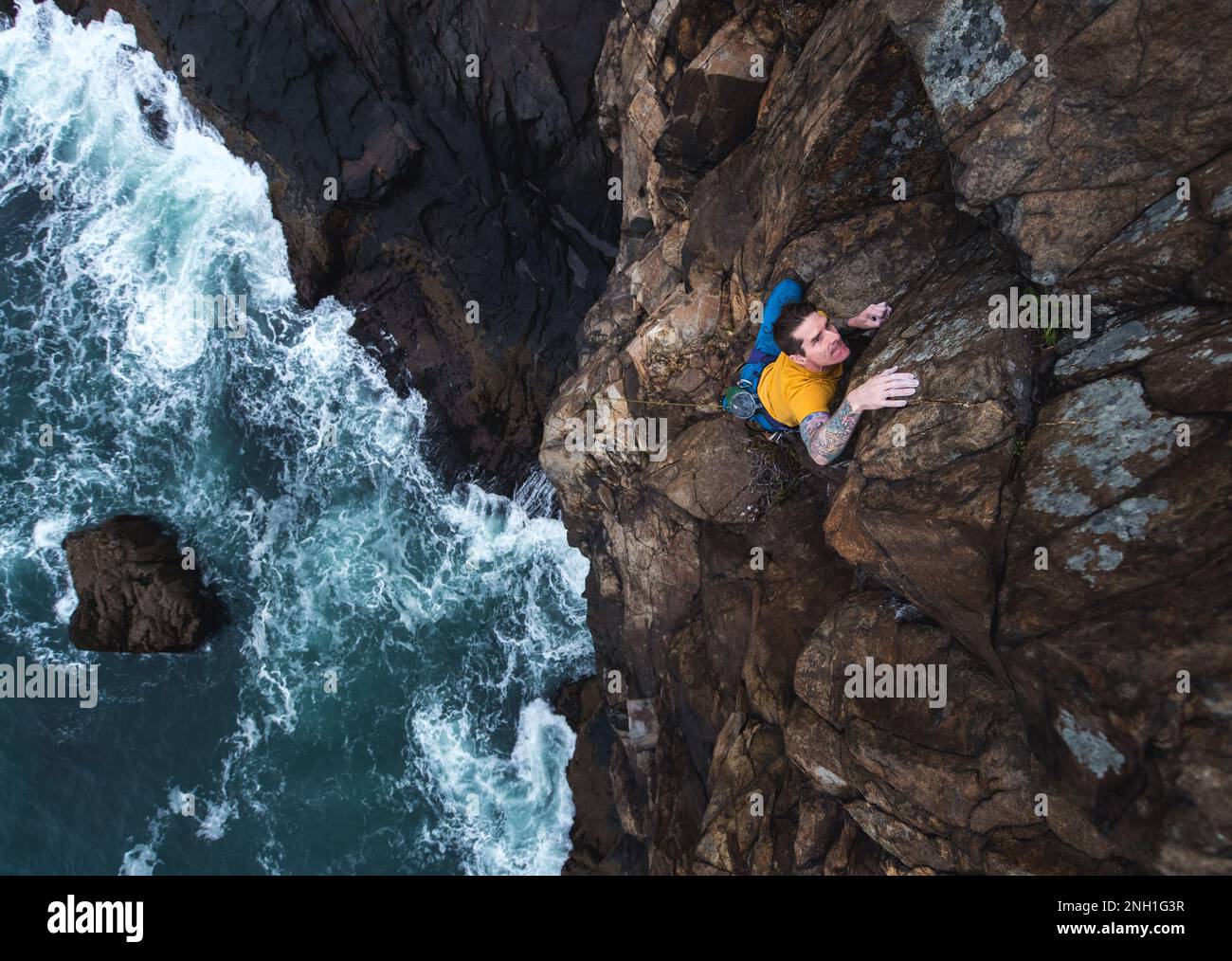 Ein Mann klettert auf eine Klippe am Meer und die Wellen schlagen darunter Stockfoto