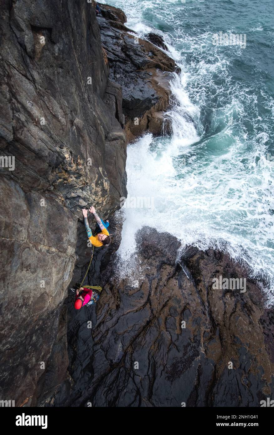 Ein Mann klettert auf eine Klippe am Meer und die Wellen schlagen darunter Stockfoto